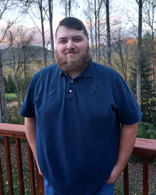 Eddie Davenport is smiling in front of a woodsy area. He is wearing a navy polo shirt, has short brown hair, and a medium length beard.